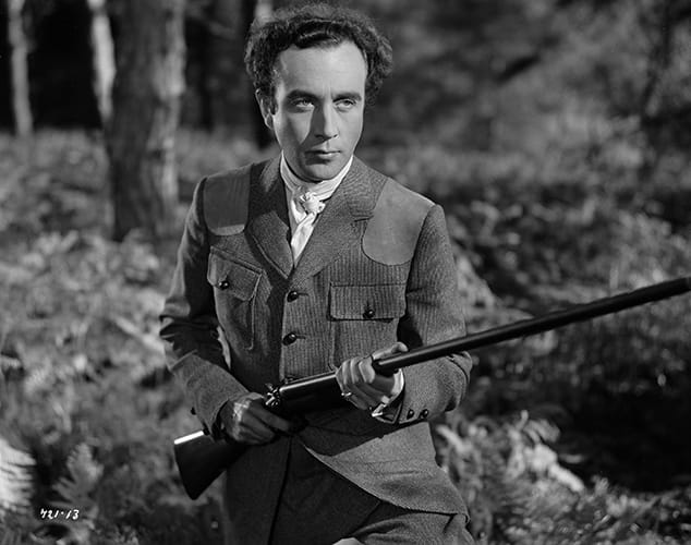 Still from Kind Hearts and Coronets showing Dennis Price in the forest holding a hunting rifle.