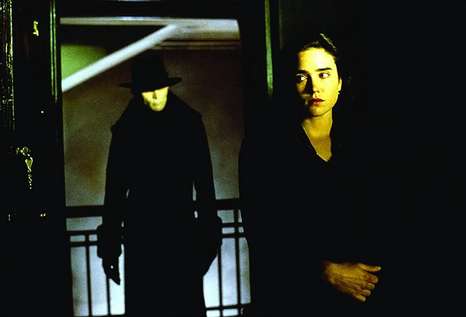 Still from Dark City showing Jennifer Connolly dressed in black, looking back over her shoulder to where a creepy man in a black coat and fedora is standing outside the window.