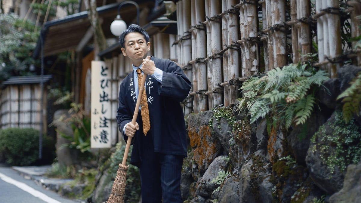 Still from River showing a Japanese man standing in the street holding a straw broom.