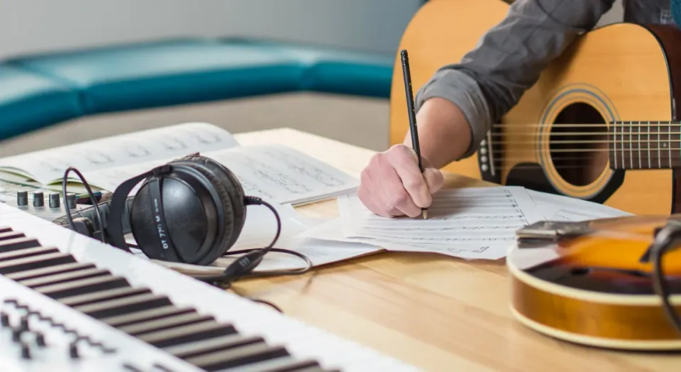 Someone sitting at a table writing on some sheet music with a guitar on their lap