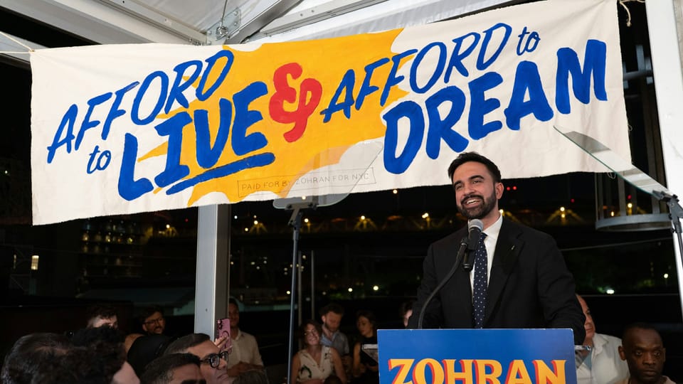 Zohran Mamdani stands at a podium in front of a banner reading "Afford to live & afford to dream"
